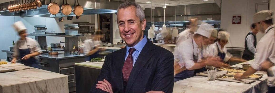 Danny Meyer standing in a restaurant kitchen with chefs working behind him, representing leadership in modern hospitality.