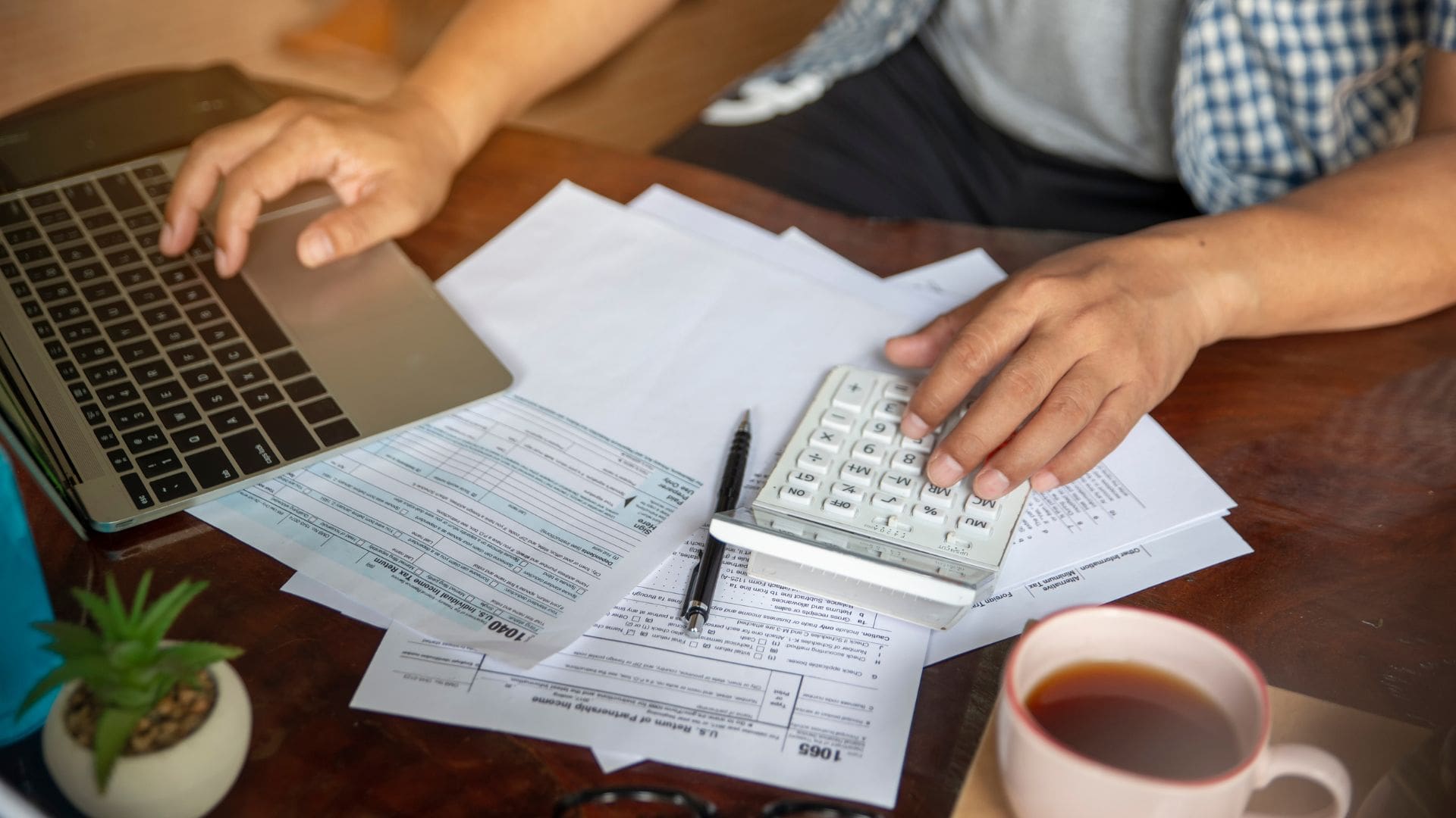 Person reviewing financial documents at a desk, using a calculator and laptop to manage expenses and budget planning.
