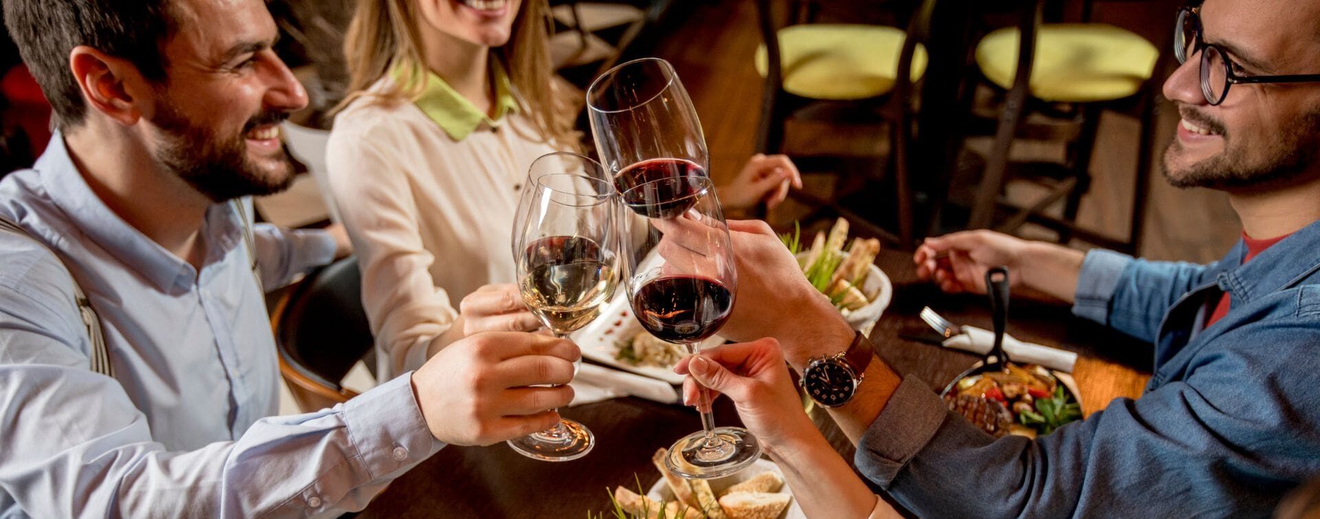 Group of friends toasting wine glasses over dinner at a restaurant table with shared plates and warm lighting.