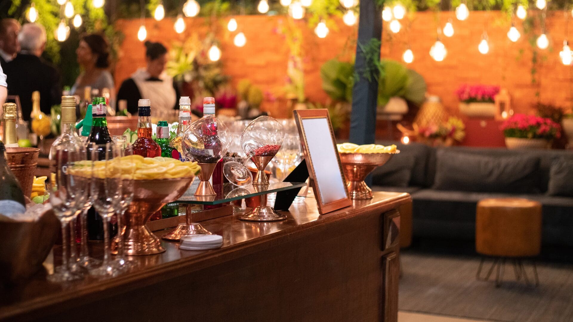 Elegant event bar setup with liquor bottles, champagne glasses, citrus garnishes, and copper barware under warm string lighting.