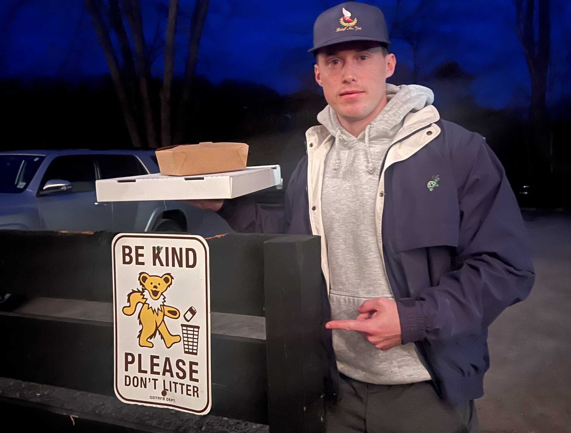 Man holding takeout containers and pointing to a “Be Kind Please Don’t Litter” sign outdoors at night, representing accountability and doing things the right way even when no one is watching