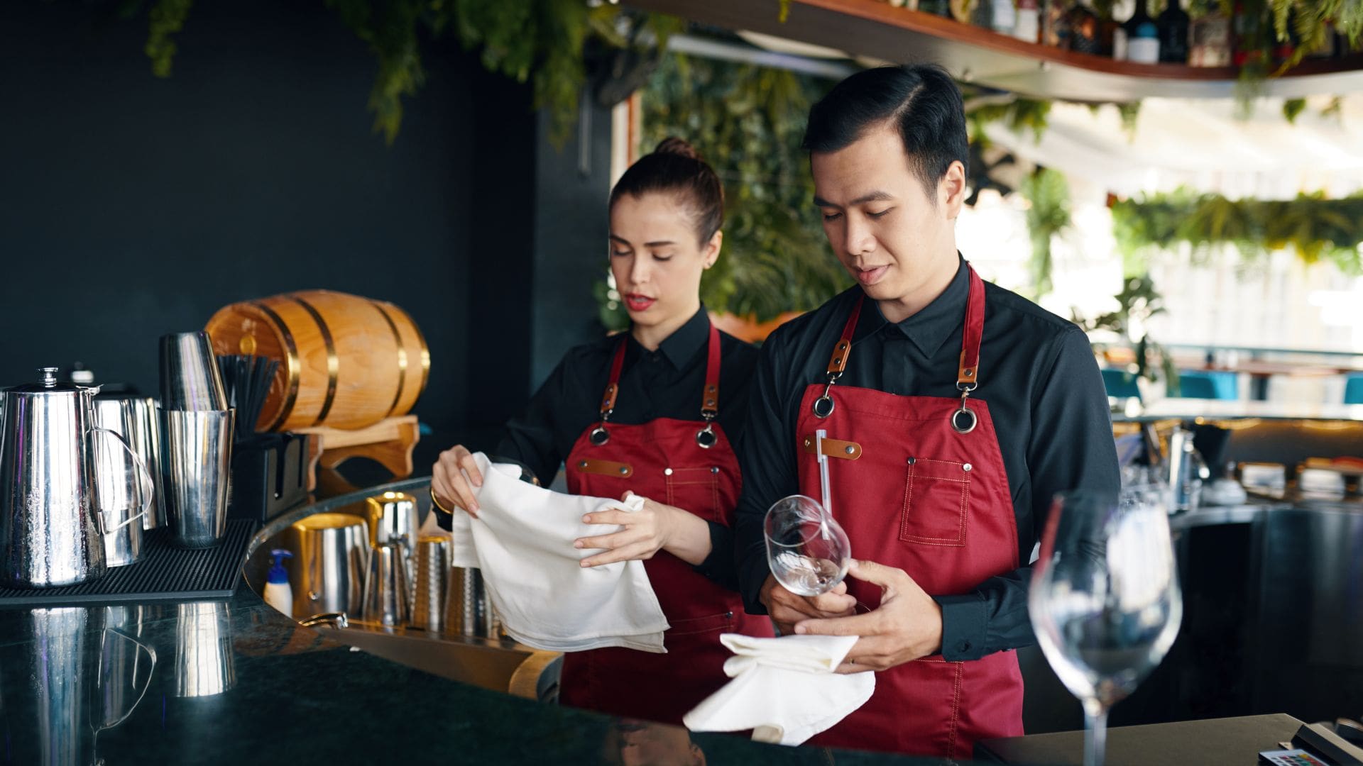 Two bartenders in red aprons polishing glassware behind a restaurant bar before a private event